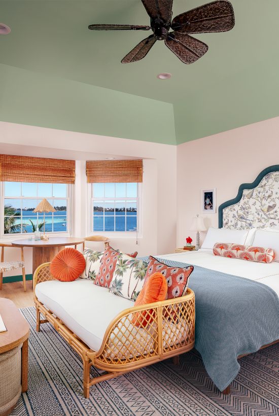 A brightly lit bedroom with a bed, chairs, a desk, and ocean view