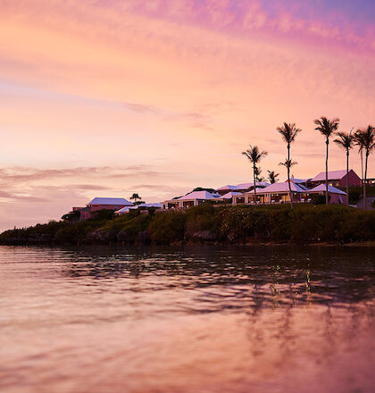 A serene sunset over a coastal area with palm trees, houses, and a calm sea reflecting the colorful sky.