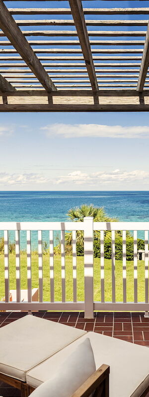 A balcony with outdoor seating overlooks a lush green lawn and a beautiful ocean view, with a partially wooden-roofed pergola overhead.