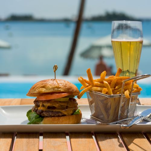A burger, fries in a metal basket, and a glass of beer on a white plate with a pool and beach in the background are shown.