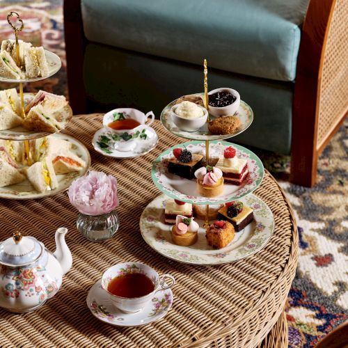 A wicker table with tea set, sandwiches, pastries, and a teapot is ready for an afternoon tea, set next to a cozy armchair.