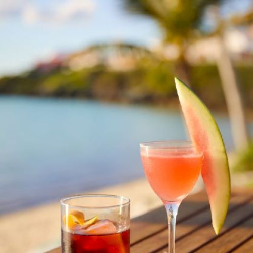 Two cocktails are on a wooden table by the beach, one garnished with an orange slice and the other with a watermelon slice.