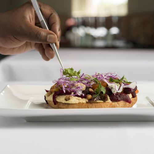 A person is plating a gourmet dish with tweezers, adding garnishes on a piece of toast topped with various colorful ingredients, on a white plate.