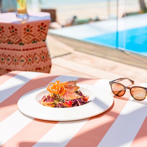 A plate of food, sunglasses, and a patterned stool are placed on a striped table near a poolside setting with a tropical vibe.