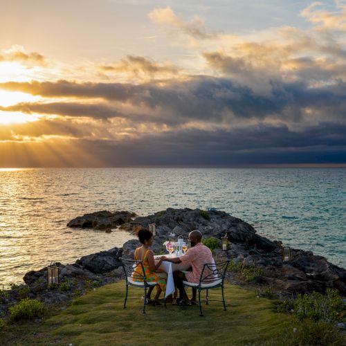 Two people sit at a table on a grassy cliff by the ocean, enjoying a meal while the sun sets in the background.