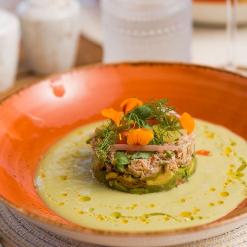 A gourmet dish featuring a layered stack of ingredients garnished with herbs and edible flowers on a creamy sauce, served in an orange bowl.