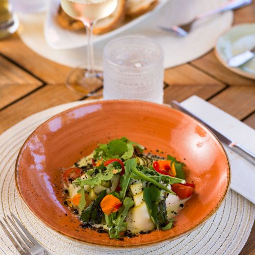 A dish of vegetables and greens on an orange plate, set on a table with a glass of white wine, cutlery, and other dining items.