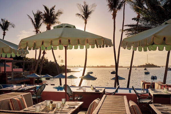 A seaside restaurant with tables, umbrellas, and palm trees overlooks a calm ocean during sunset, creating a tranquil atmosphere.