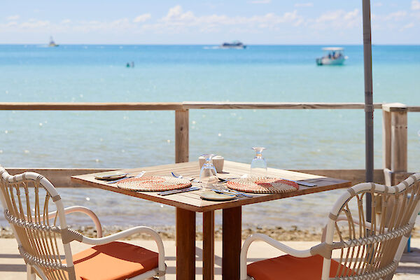 A seaside café setup featuring a table with two chairs under a large umbrella, overlooking calm ocean waters with boats in the distance.