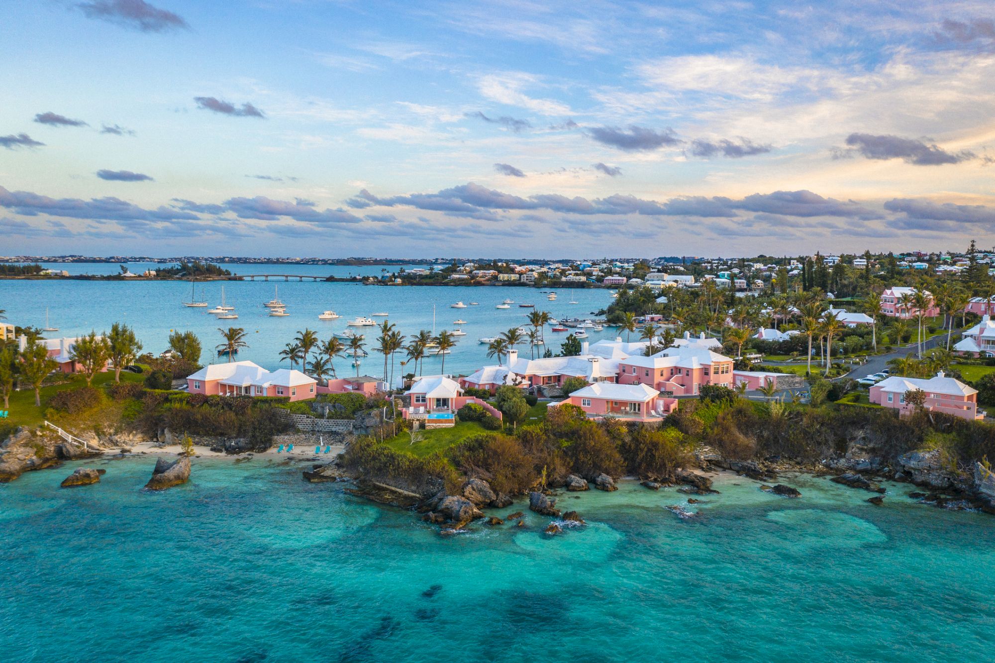 A coastal scene features turquoise waters, pink and white houses, palm trees, and boats in a harbor with a partly cloudy sky.