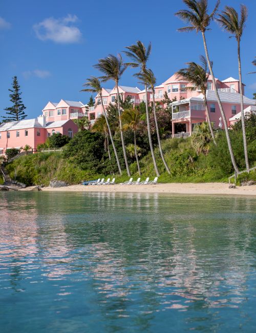 The image shows pastel-colored buildings on a hillside with palm trees, overlooking a calm beach and clear blue water under a bright sky.