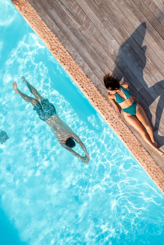 Two people are by a pool, one is swimming while the other is lounging on a wooden deck beside the water, casting shadows.