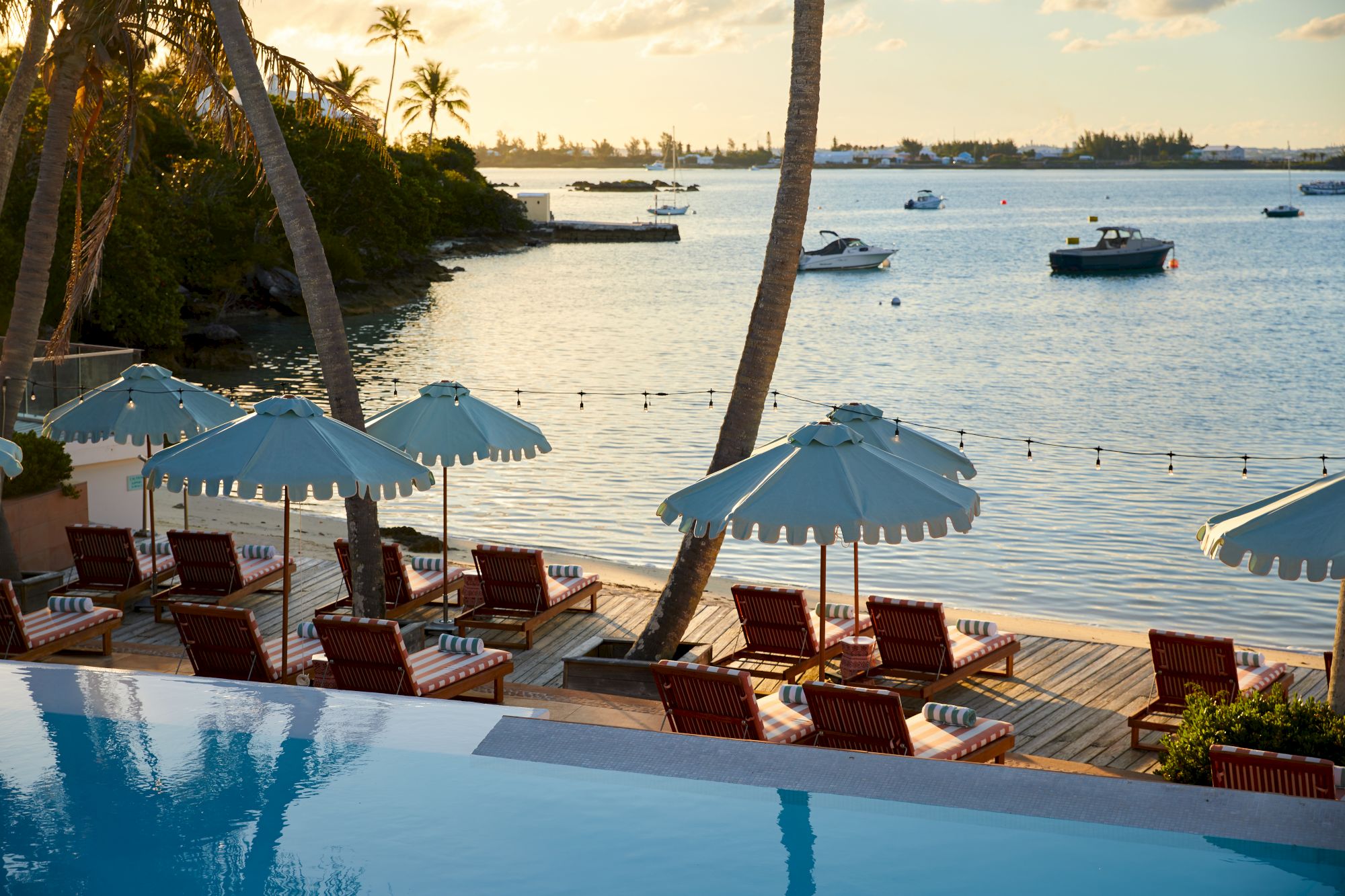 A serene waterfront scene featuring lounge chairs, umbrellas, an infinity pool, and moored boats, with palm trees and sunset in the background.