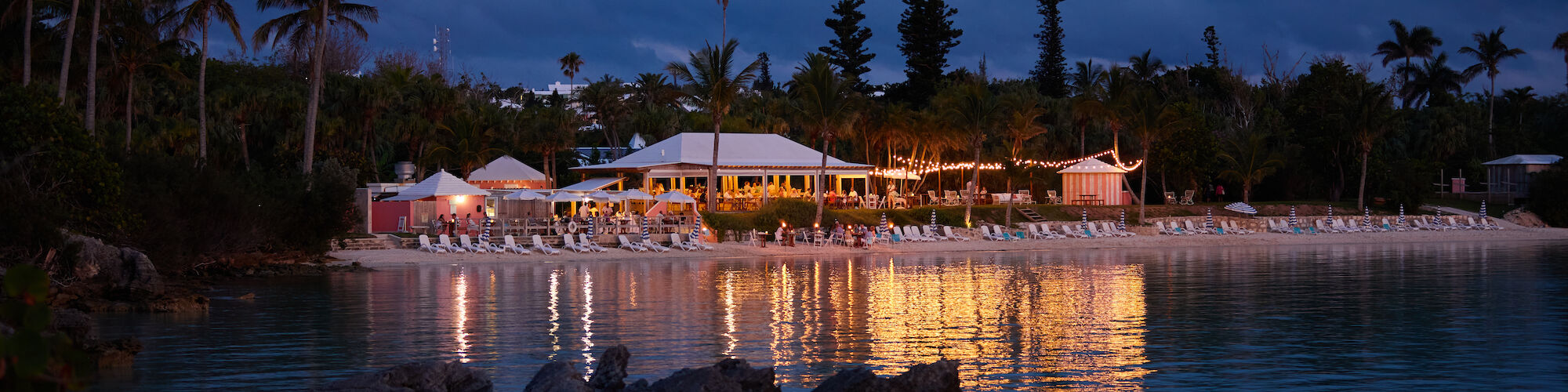 A serene beach scene at dusk with lit buildings, palm trees, and reflections on the calm water. Rocks are partially submerged in the foreground.