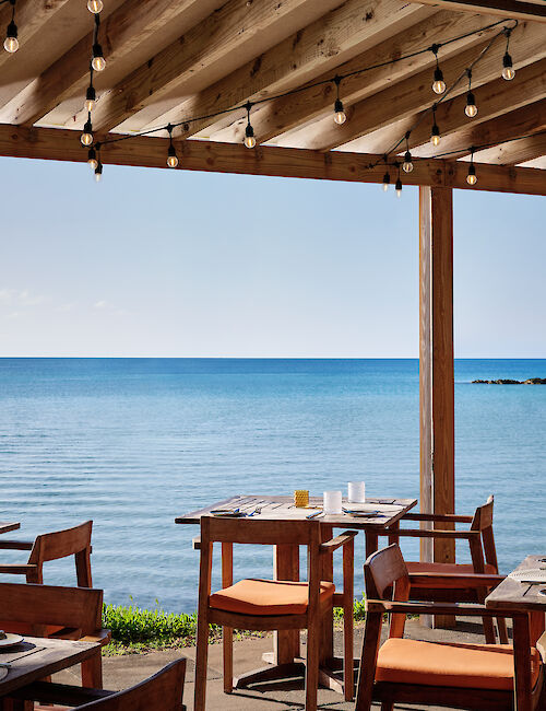 The image shows an outdoor restaurant with wooden tables and chairs overlooking a calm, blue ocean under a wooden canopy with string lights.