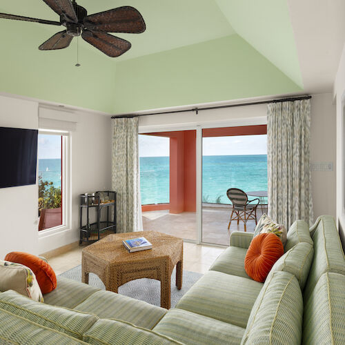 A living room with a green striped sofa, wicker coffee table, flat-screen TV, ceiling fan, and view of the ocean from a balcony with chairs.