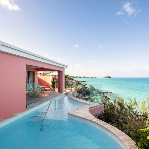 A seaside pink building with a pool, overlooking clear blue waters and surrounded by greenery under a bright sky.