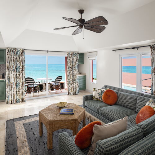 A cozy living room with beach decor, featuring a gray sofa, colorful pillows, a ceiling fan, and large windows offering a view of the ocean.