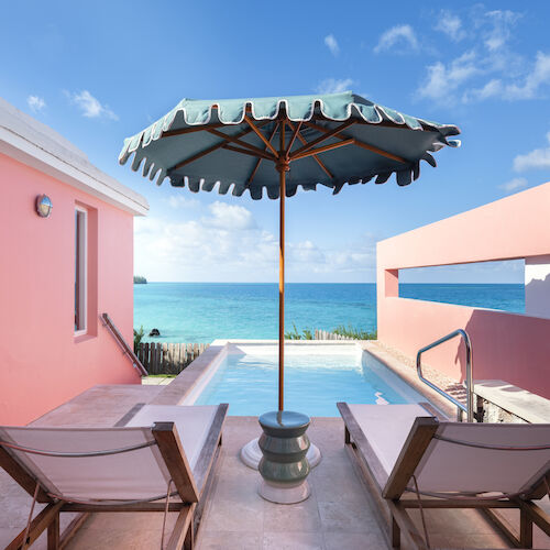The image shows a seaside terrace with two lounge chairs under an umbrella, a pool, and a view of the ocean, framed by pink walls.