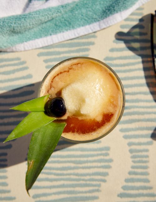 The image shows a tropical cocktail garnished with a cherry and pineapple leaves, placed on a patterned surface near a pair of sunglasses and a towel.