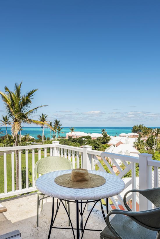 A scenic beach view from a balcony with a table, chairs, and a hat on the table, overlooking palm trees, blue ocean, and houses in the distance.