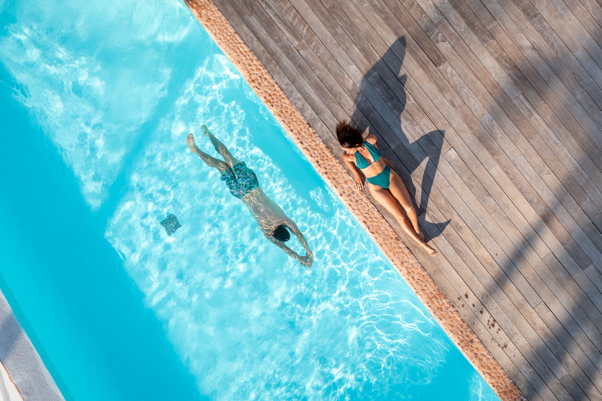 A person swims in a pool while another person relaxes on a wooden deck beside it, casting a shadow in the shape of a shark.