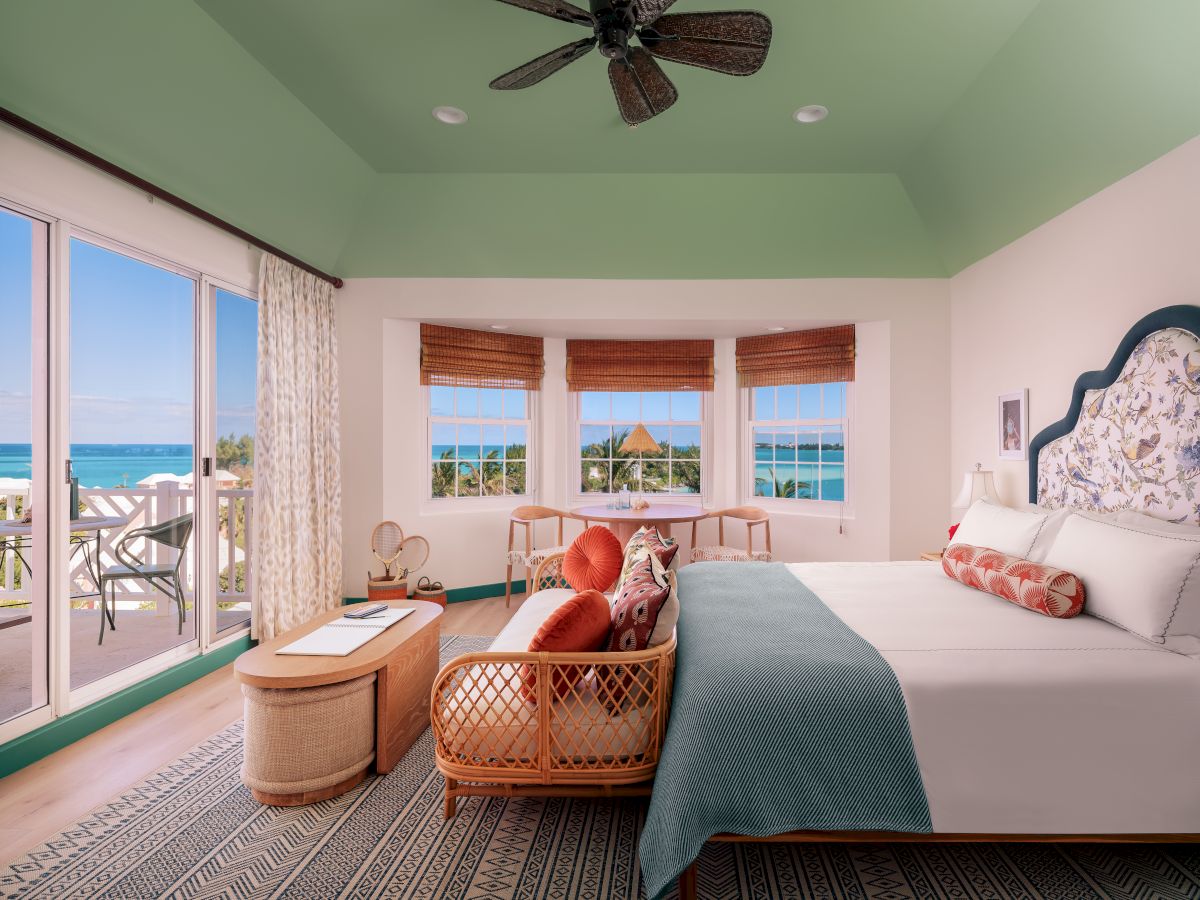 A bright bedroom with ocean views features a large bed, wicker sofa, coffee table, large windows, sliding doors, and a ceiling fan, ending the sentence.