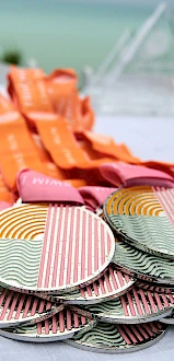 The image shows a collection of medals with colorful, striped designs and orange ribbons, arranged on a table near what appears to be a beach.