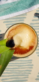 A tropical cocktail garnished with pineapple leaves and a cherry is on a patterned surface, beside a towel and sunglasses.