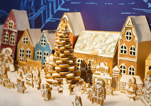 A festive gingerbread village with colorful houses, a gingerbread tree, and various gingerbread figures is set on a snowy base.