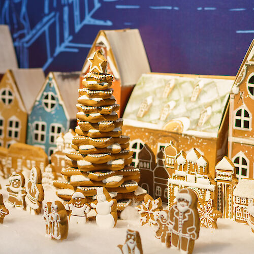 A festive gingerbread village with colorful houses, a gingerbread tree, and various gingerbread figures is set on a snowy base.