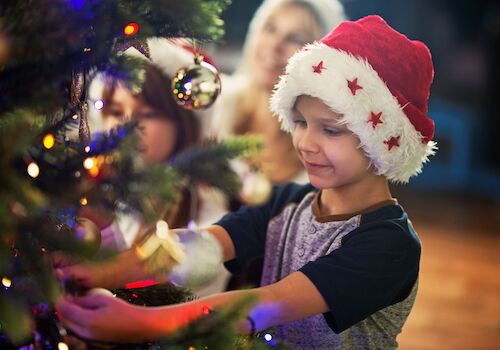 A child in a Santa hat decorates a Christmas tree, with two people in the background. The scene is festive and joyful.