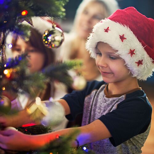 A child in a Santa hat decorates a Christmas tree, with two people in the background. The scene is festive and joyful.