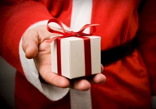 A hand in a Santa Claus costume holding a small, white gift box with a red ribbon tied in a bow.