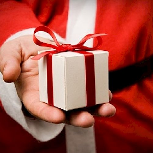 A hand in a Santa Claus costume holding a small, white gift box with a red ribbon tied in a bow.