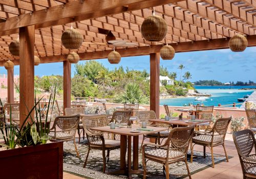 An outdoor dining area with wooden furniture, wicker chairs, hanging lamps, and a view of the ocean and greenery in the background.