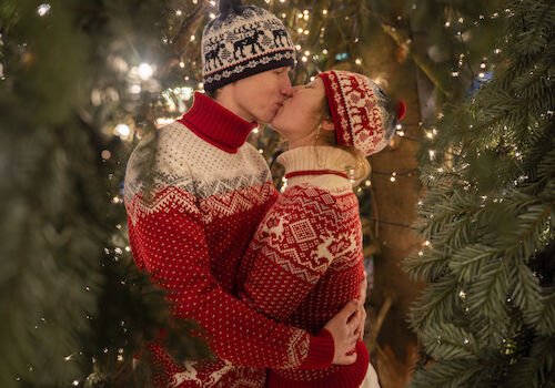 A couple kissing among Christmas lights, wearing festive sweaters and beanies, surrounded by greenery and a cozy holiday atmosphere.