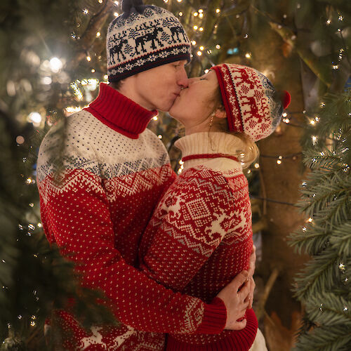 A couple kissing among Christmas lights, wearing festive sweaters and beanies, surrounded by greenery and a cozy holiday atmosphere.