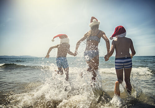 Three people in swimwear and Santa hats joyfully run into the ocean.