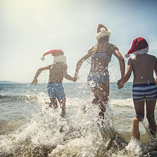 Three people in swimwear and Santa hats joyfully run into the ocean.