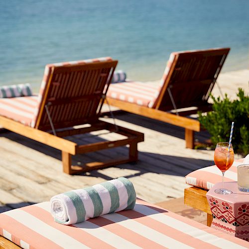 Two wooden sun loungers with pink-striped cushions and rolled towels are on a wooden deck near the water, featuring two colorful drinks on a table.