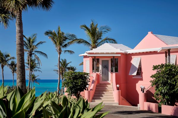 A pink house with white trim and surrounded by palm trees sits near a vibrant blue ocean under a clear sky, creating a tropical setting.
