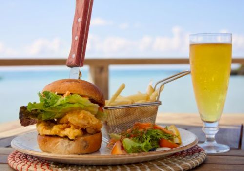 A burger with a knife, fries in a basket, a salad, and a glass of beer are served on a plate with a scenic view of the ocean in the background.