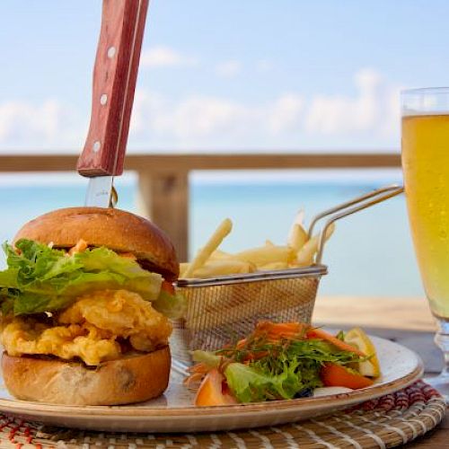 A burger with a knife, fries in a basket, a salad, and a glass of beer are served on a plate with a scenic view of the ocean in the background.