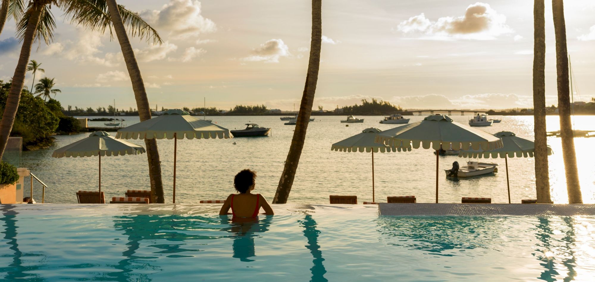 A person relaxes in a pool overlooking a scenic waterfront with boats, parasols, and palm trees at sunset.