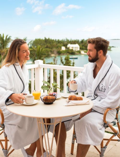 Two people in robes enjoy breakfast at an outdoor table on a balcony with a scenic view of water and lush greenery in the background.