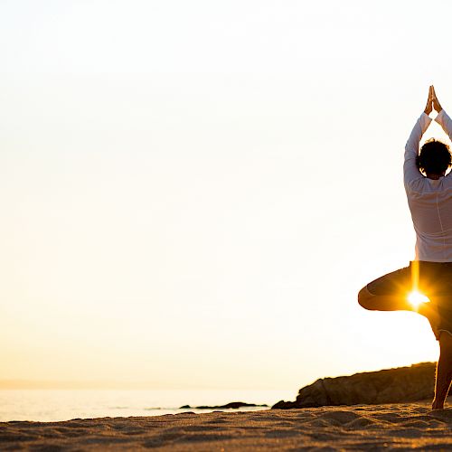 A person is doing a yoga pose on the beach at sunset, with the sun shining through their legs, creating a serene and peaceful setting.