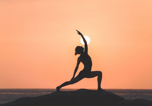 A person is performing a yoga pose on a rock during sunset, with the sun directly behind them, creating a serene silhouette.