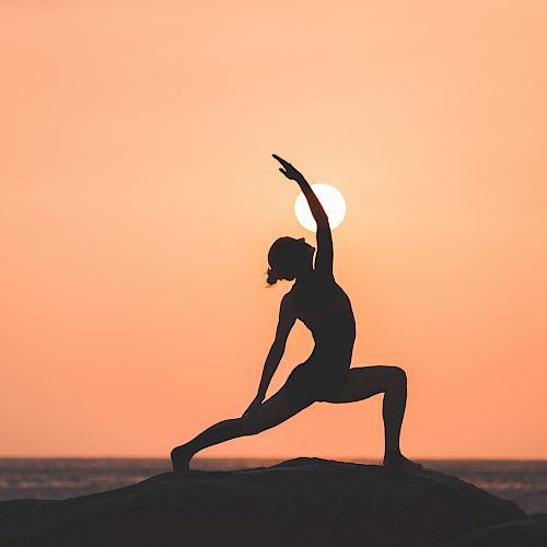 A person is performing a yoga pose on a rock during sunset, with the sun directly behind them, creating a serene silhouette.