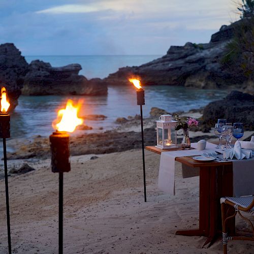 A romantic beachside dinner setup with a table for two, surrounded by lit torches and rocky landscape by the ocean at dusk, creating a serene ambiance.